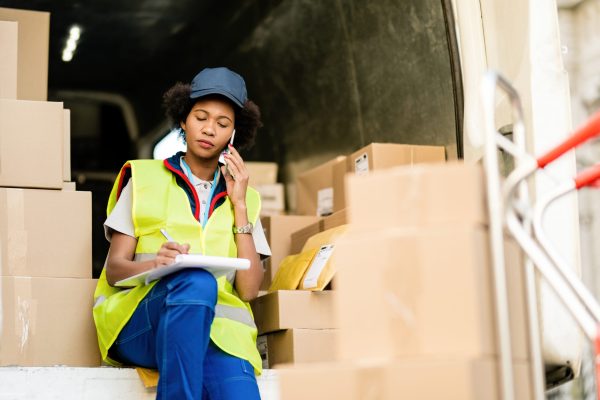 Female black courier sitting in delivery van and communicating on mobile phone while writing notes in clipboard.